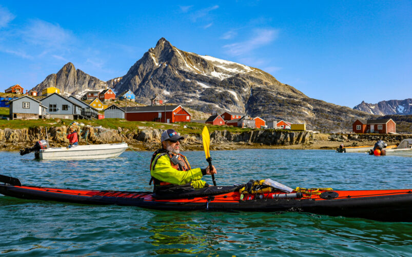 Voyage polaire à Kulusuk, kayak de mer devant un village coloré du Groenland avec montagnes escarpées en arrière-plan.