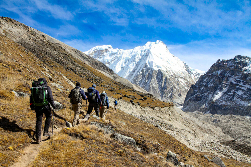 Un groupe de randonneurs emprunte un étroit sentier de montagne entouré d'un terrain rocailleux, avec des sommets enneigés comme le Makalu au loin et un ciel bleu vif au-dessus, capturant l'esprit d'une traversée de l'Himalaya à couper le souffle.