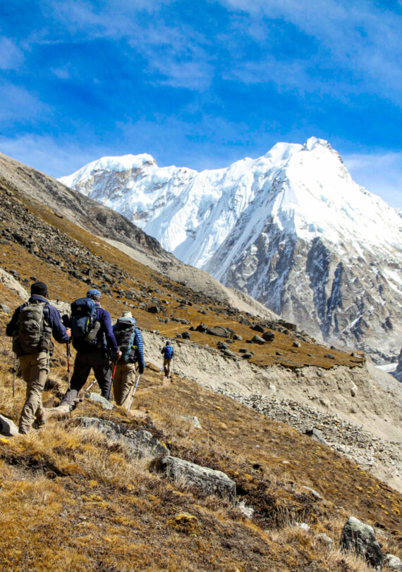 Un groupe de randonneurs emprunte un étroit sentier de montagne entouré d'un terrain rocailleux, avec des sommets enneigés comme le Makalu au loin et un ciel bleu vif au-dessus, capturant l'esprit d'une traversée de l'Himalaya à couper le souffle.