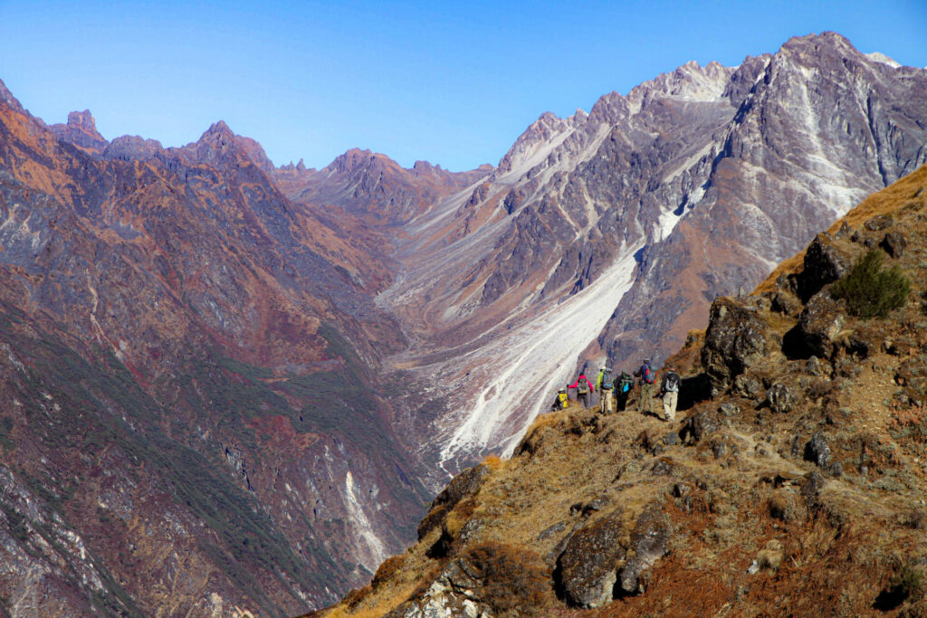 Un groupe de randonneurs se tient sur une crête rocheuse surplombant de vastes pentes de montagnes brunes et violettes près du Kangchenjunga, avec des pics enneigés sous un ciel bleu clair.